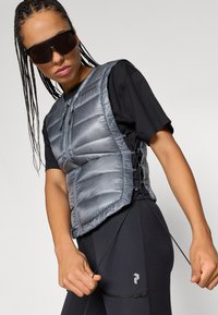 Pūkuotas pilkas liemenė su užtrauktuku priekyje ir šoninėmis juostelėmis, apsirengus juodą marškinį, derinamas su tamsiais saulės akinių akiniais. Juodi legginsai turi šoninės kišenės detalę.