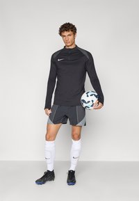 Svart långärmad träningströja med grå axelaccenter, grå shorts med geometriska mönster, vita strumpor och svarta fotbollsskor. Håller i en fotboll.