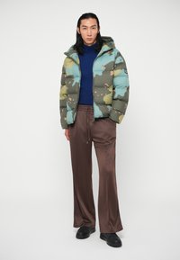 Seisev inimene, kellel on seljas sinine kõrge kaelusega jakk, kamoflaažiga puhvrjakker, laiad pruunid püksid ja mustad kingad, taustal ühtlane valge taust.