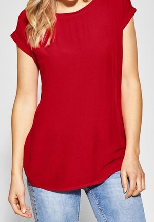 Frau trägt ein einfarbiges rotes Kurzarmshirt und hellblaue Jeans, steht mit entspannten Armen seitlich am Körper.