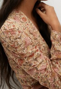 T-shirt à manches longues en tissu beige clair avec un imprimé floral en rose, rouge et marron. Détails froncés aux épaules ajoutent de la texture.