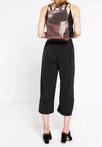 Sac à dos en or rose métallique avec texture à motif, forme rectangulaire, fermeture à rabat, porté dans le dos ; assorti à un pantalon court à fines rayures noires.