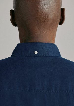 Back of a bald person's head wearing a dark blue shirt with a button at the center of the collar.