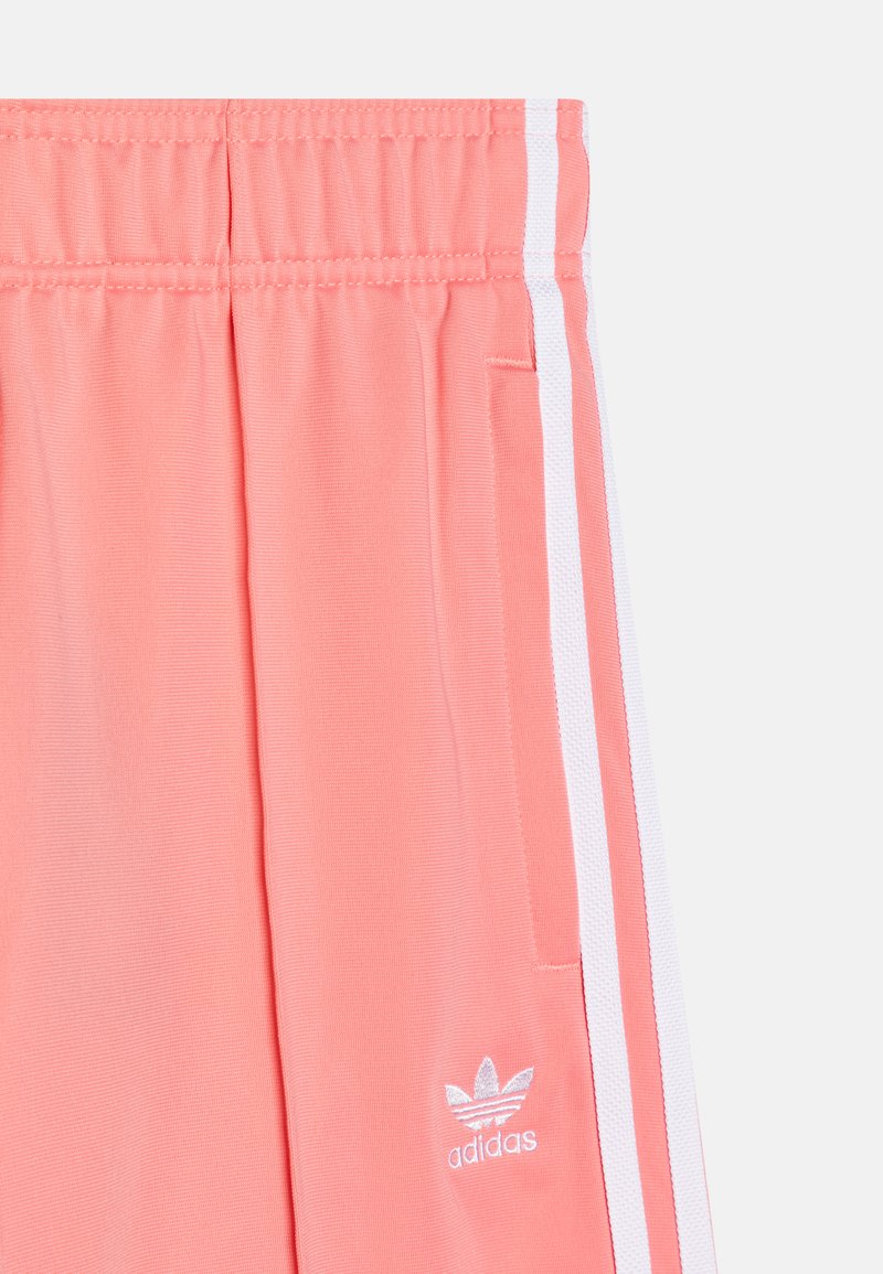 Różowe szorty sportowe z białymi bocznymi paskami, elastycznym pasem i małym białym logo Adidas na dolnej części nogawki. Gładki, lekki materiał.