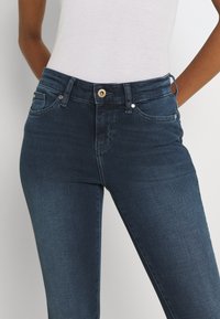 Jeans i mörkt indigo, med en figurnära design, fem fickor och en guldknappstängning. Slät textur med lätt stretch.