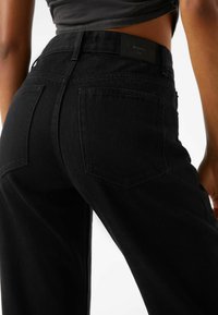 Jeans de mezclilla negra con cintura ajustada, dos bolsillos en la parte posterior y una etiqueta de logotipo bordada en la cinturilla. Tejido texturizado con un acabado suave.