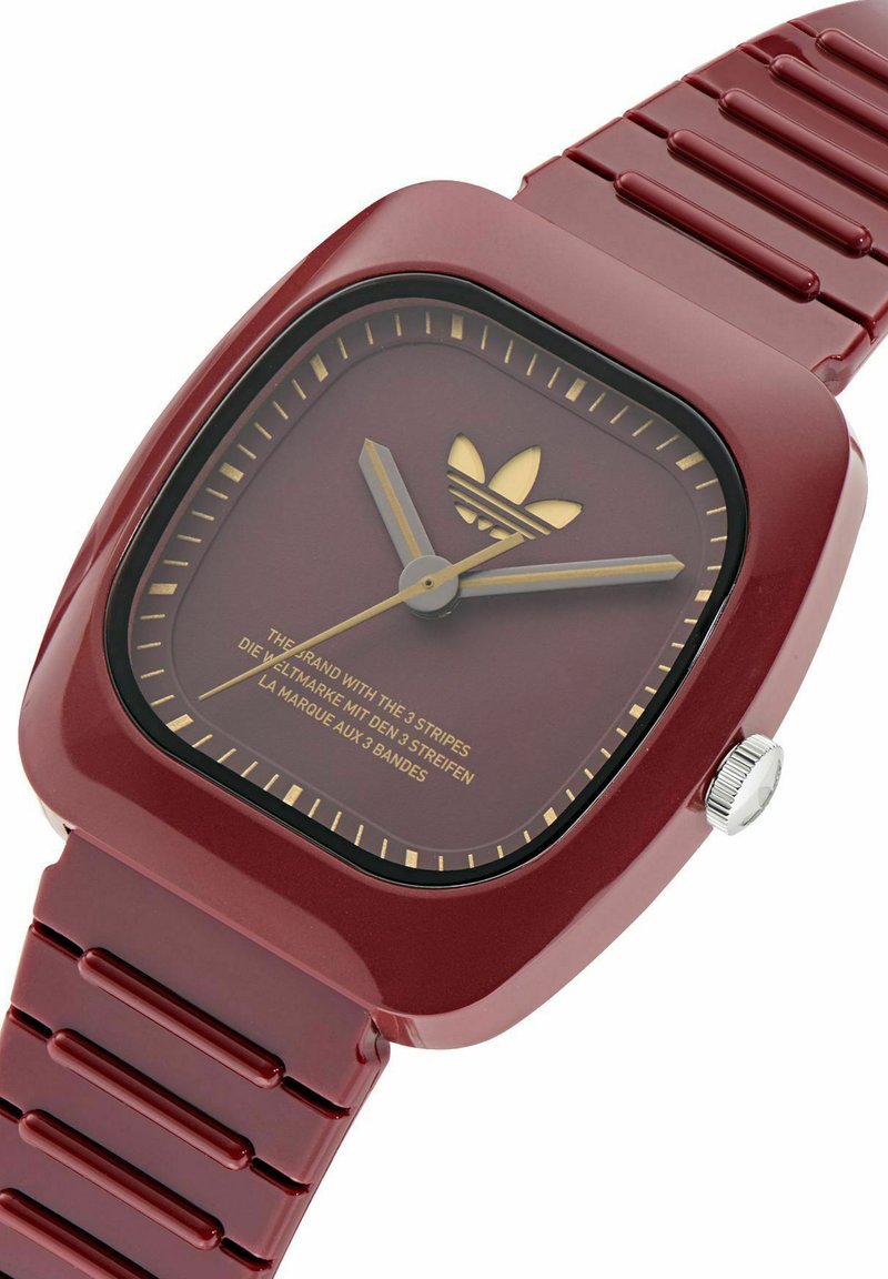 Orologio quadrato rosso con cinturino in gomma, dettagli dorati, quadrante minimalista e logo. Presenta una corona sul lato e maglie del cinturino testurizzate.