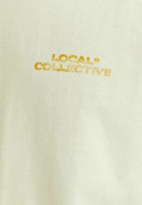 Šviesiai žalia medvilninė marškinėliai, su įspūdingu auksiniu spausdintu tekstu „LOCAL + COLLECTIVE“, turinčiu šiek tiek tekstūrinį vaizdą.