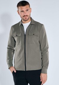 Olivgrüne Jacke mit Reißverschluss, ausgestattet mit zwei Brusttaschen, langen Ärmeln und einer glatten Textur. Kombiniert mit einem weißen T-Shirt darunter.