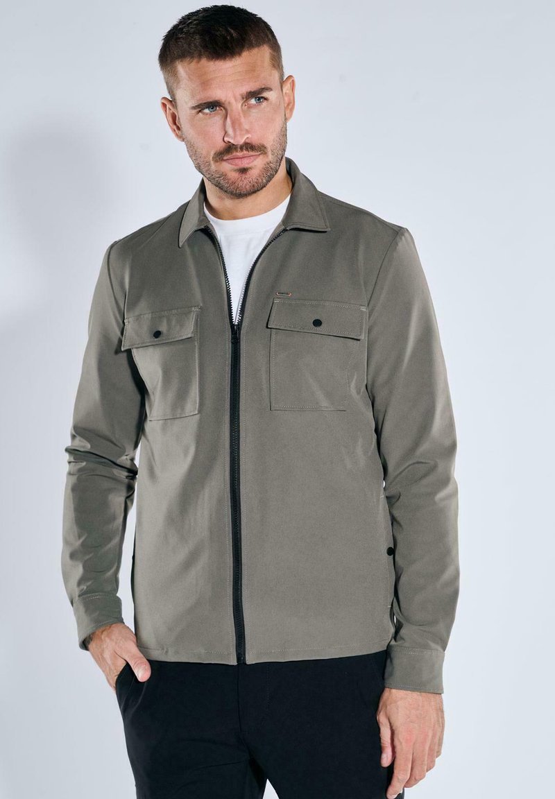 Olivgrüne Jacke mit Reißverschluss, ausgestattet mit zwei Brusttaschen, langen Ärmeln und einer glatten Textur. Kombiniert mit einem weißen T-Shirt darunter.