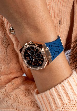 Reloj Guess con caja de oro rosa, esfera negra, números en negrita, detalles de cristal y pulsera de silicona azul texturizada, llevado en una muñeca.