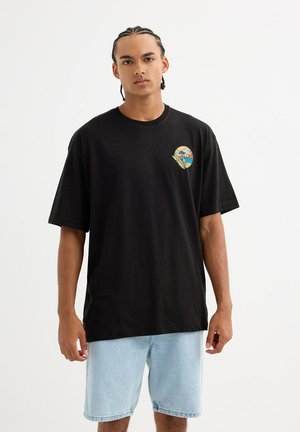 Camiseta de algodón negra con un parche gráfico redondo que presenta un diseño de esqueleto sobre un fondo colorido. Mangas de ajuste estándar.