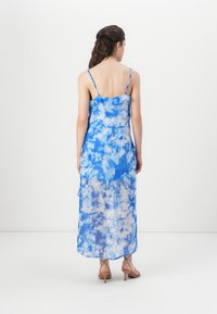 Μακρύ φόρεμα tie-dye σε μπλε απόχρωση με λεπτές τιράντες, ελαφρύ ύφασμα και ρέουσα σιλουέτα. Ο σχεδιασμός διαθέτει έναν συνδυασμό ανοιχτόχρωμων και σκούρων μπλε αποχρώσεων.