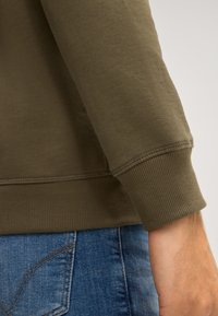 Oliwkowy sweter z ribbami przy mankietach i dolnej krawędzi, charakteryzujący się gładką teksturą i dopasowanym krojem, zestawiony z niebieskimi dżinsami.