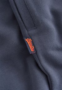 Tela azul marino con costuras visibles y una pequeña etiqueta oscura con texto estilizado en naranja brillante en la costura.