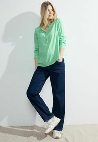 Camicia button-up a righe verde chiaro, vestibilità rilassata; abbinata a jeans blu scuro e sneakers bianche; appoggiata su uno sfondo semplice.