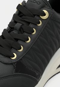 Black sneaker with quilted side panels, black laces threaded through gold eyelets, and white sole detail.
