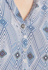 Camicia a motivo geometrico blu e bianco con quattro bottoni decorativi argentati, che mostra una parte del collo e dell'area del colletto.