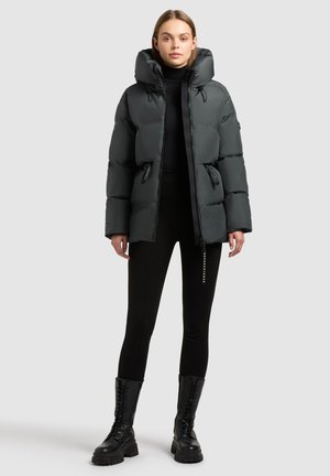 Giacca a vento nera con colletto alto, chiusura con zip e due tasche laterali. Indossata sopra un top nero e leggings, abbinata a stivali neri con lacci.