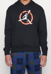 Persona con sudadera negra y el logo de Air Jordan Flight Club en rojo y blanco, con pantalones de cuadros azul-verde-rojo, de pie contra un fondo liso.