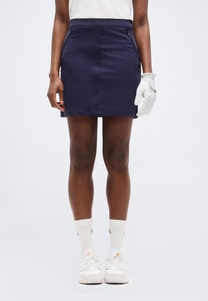 Oseba, oblečena v mornarsko modro golf krilo, z belim rokavico na desni roki, belimi nogavicami in belimi supergami na enobarvnem ozadju.