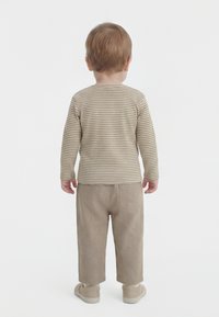 Malček stoji obrnjeno stran, oblečen v bež črtasto majico z dolgimi rokavi, bež hlače in bež čevlje na belem ozadju.