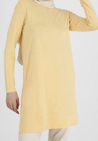 Nainen, joka pukeutuneena pitkähiharaiseen vaaleankeltaiseen tunikaan vaaleiden housujen kanssa, ja kaulassa vaaleanvärinen huivi, poseeraa yksi käsi nostettuna.