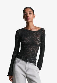 Top lungo in pizzo nero con maniche lunghe e scollatura a barca, caratterizzato da un pattern testurizzato e un orlo ondulato, abbinato a jeans grigi chiari.