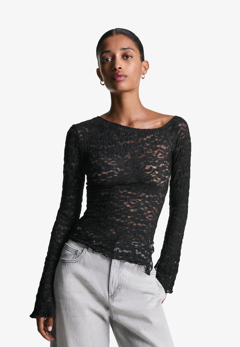 Top lungo in pizzo nero con maniche lunghe e scollatura a barca, caratterizzato da un pattern testurizzato e un orlo ondulato, abbinato a jeans grigi chiari.