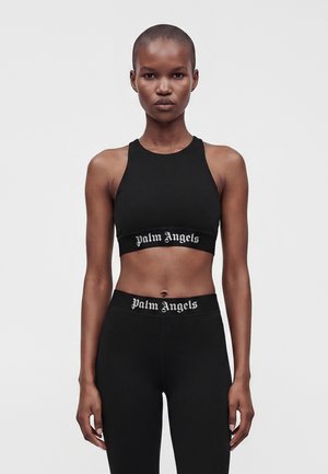 Γυναίκα που φοράει μαύρο αμάνικο crop top και κολάν με λευκό κείμενο ""Palm Angels"" στις ζώνες, στέκεται μπροστά σε απλό φωτεινό φόντο.
