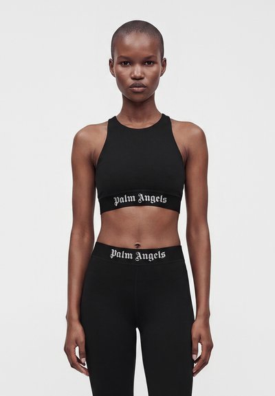 Γυναίκα που φοράει μαύρο αμάνικο crop top και κολάν με λευκό κείμενο ""Palm Angels"" στις ζώνες, στέκεται μπροστά σε απλό φωτεινό φόντο.