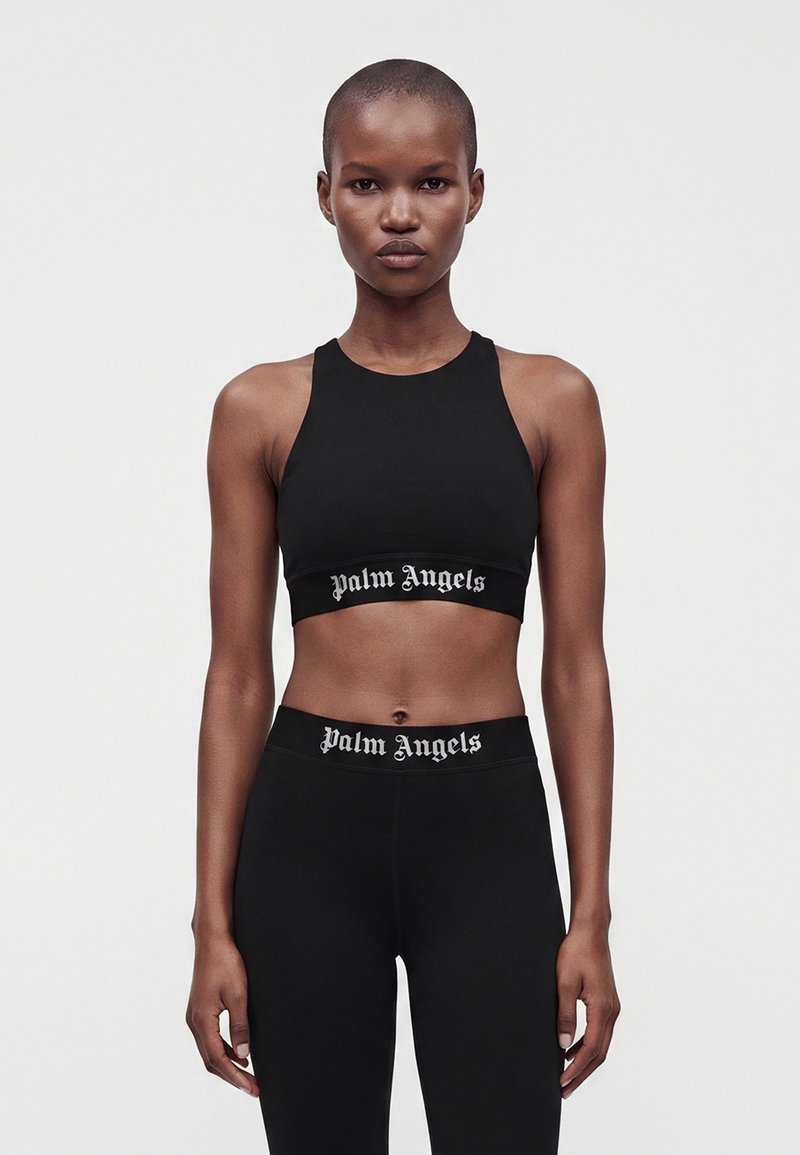 Γυναίκα που φοράει μαύρο αμάνικο crop top και κολάν με λευκό κείμενο ""Palm Angels"" στις ζώνες, στέκεται μπροστά σε απλό φωτεινό φόντο.