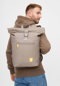 Grauer Rolltop-Rucksack mit verstellbaren Trägern, großem Reißverschlussfach, gummiertem Material, gelbem Logo-Patch, schlankem Design.