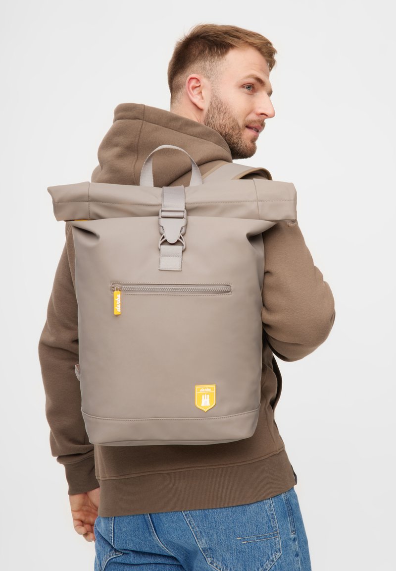 Grauer Rolltop-Rucksack mit verstellbaren Trägern, großem Reißverschlussfach, gummiertem Material, gelbem Logo-Patch, schlankem Design.