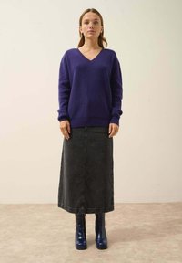 Lila v-ringad tröja med lös passform och ribbade ärmmuddar, parat med en lång svart denimkjol och blå klackstövlar, stående mot en neutral bakgrund.