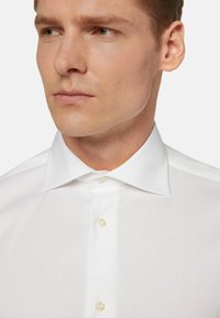 La camicia bianca presenta un colletto classico, una chiusura anteriore con bottoni color crema e un tessuto liscio e strutturato. Primo piano della parte superiore del corpo.