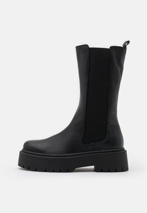 Botte de cheville noire avec une tige en cuir lisse, un panneau latéral élastique et une semelle en caoutchouc côtelée et chunky pour une meilleure traction.