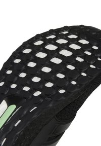 Semelle de chaussure de sport noire avec caoutchouc texturé présentant des motifs circulaires et rectangulaires, semelle intermédiaire blanche et un petit accent vert.
