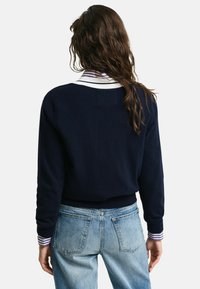 Maglione blu navy con colletto bianco a righe, maniche lunghe e vita aderente, indossato sopra jeans di denim azzurri.