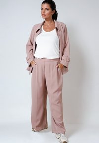 Blush-pink maßgeschneiderte Blazerjacke und plissierte Hose, kombiniert mit einem weißen, gerippten Tanktop. Das Outfit umfasst Taschen und lässige Sneakers.