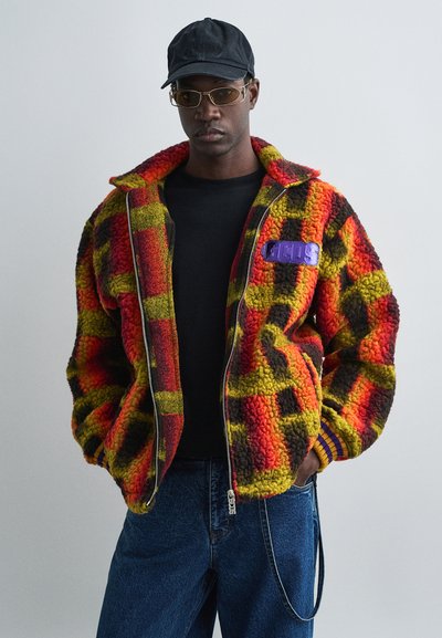 Veste en polaire avec un motif écossais rouge, jaune et noir ; fermeture éclair, poignets côtelés et un patch logo violet sur la poitrine.