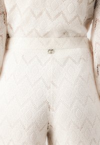Outfit bianco in pizzo con un motivo geometrico e una finitura texturizzata. Il dettaglio sul retro include un piccolo gancio metallico in vita.
