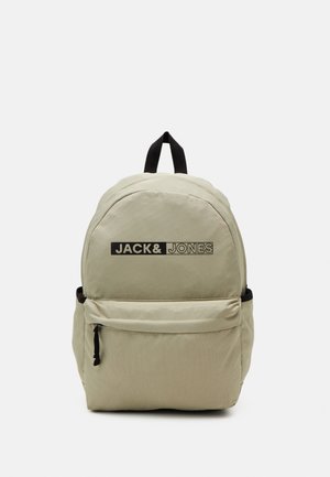 Zaino beige dalla forma arrotondata, con una tasca frontale, dettagli neri e un logo stampato in bianco e nero sulla parte anteriore.