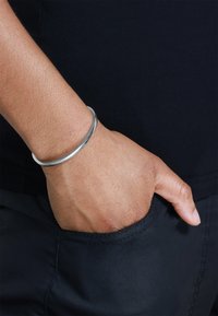 Sølv armbånd på en hånd, glatt overflate og sirkulær form, båret over en mørk lomme på et plagg. Hud med en lys tekstur er synlig.