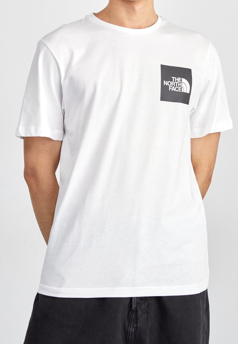 Vit bomullströja med rund halsringning, korta ärmar och en svart fyrkantig ficka med "The North Face"-logotypen i vit text.
