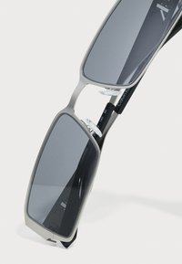 Ochelari de soare cu un cadru metalic argintiu, design rectangular, lentile cu nuanța gri și dopuri nazale ajustabile; brațe negre cu detalii ale mărcii.