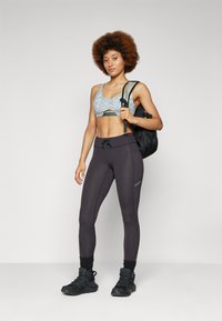 Top desportivo azul claro com um design recortado, combinado com leggings cinzas escuras com uma costura elegante. O modelo usa botas de caminhada pretas e uma mochila preta.