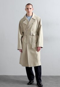 Cappotto trench beige doppiopetto con ampi revers, vita cinturata e tasche laterali. Indossato sopra una maglia azzurro chiaro, abbinato a pantaloni neri.
