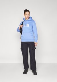 Felpa con cappuccio azzurro chiaro e logo bianco, abbinata a pantaloni cargo neri con tasche. Il modello tiene una borsa nera e indossa scarpe nere.