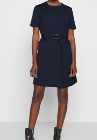 Robe en coton bleu marine avec des manches courtes, un col rond et une taille ceinturée avec une boucle en argent. Texture lisse et design évasé.